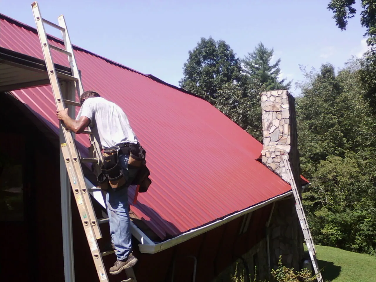Roofer on a ladder beside a red metal roof and stone chimney — Soffit & Fascia Repair by Roofing Brothers in Clarksburg